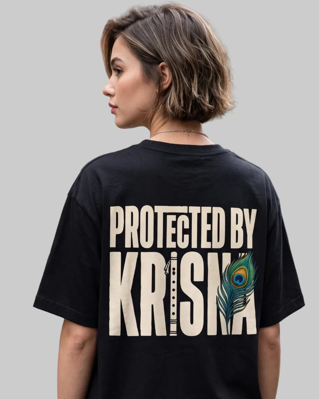 A woman wearing a black graphic t-shirt with 'PROTECTED BY KRISHNA' printed in beige on the back, standing on a cobblestone street in front of brick buildings.